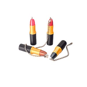 May include: Four lipstick-shaped keychains featuring a black base, a gold-colored middle section, and a colored top. Two are upright with pink and red tops, while two are lying down with metallic brown and black tops, each attached to a silver ball chain.
