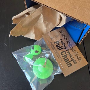 K&ouml;nnte beinhalten: Ein hellgr&uuml;ner Alien- und UFO-Deckenventilator-Zugschalter in einer durchsichtigen Plastikt&uuml;te. Ein Pappschild tr&auml;gt die Aufschrift Wooden Androyd Studio Ceiling Fan/Light Pull Chain. Der Artikel befindet sich in einem Karton aus Pappe.