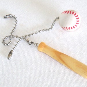 May include: A baseball-themed ceiling fan pull chain featuring a miniature baseball with red stitching and a wooden baseball bat handle. The chain is made of silver-colored metal.