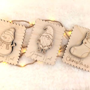 Puede incluir: Tres adornos navideños de madera con bordes festoneados. Uno presenta un muñeco de nieve, otro un gnomo y el último un calcetín. Cada uno tiene un diseño festivo y las palabras "Joyeux Noel" o "Merry Christmas" grabadas.