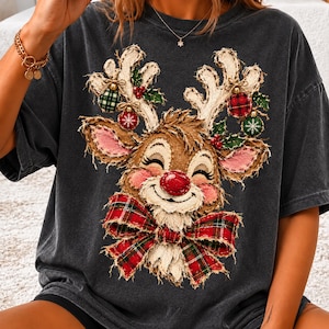 Può includere: Una t-shirt a maniche corte grigio antracite con la grafica di una renna sorridente dal naso rosso, che indossa un grande fiocco a quadri rossi e verdi. Le corna della renna sono decorate con piccoli ornamenti a quadri e foglie di agrifoglio.