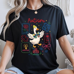 May include: Black t-shirt with a colorful graphic design featuring a goose wearing a bow tie and a yellow hat. The word "Autism" is written in red script above the goose. Surrounding the goose are puzzle pieces and the words "accept", "advocate", "educate", and "love".