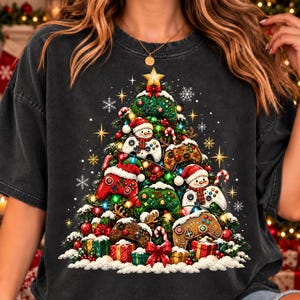 May include: A charcoal grey t-shirt featuring a graphic of a Christmas tree decorated with various video game controllers, snowmen, candy canes, and wrapped gifts, topped with a gold star and surrounded by white snowflakes.