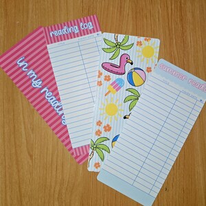 May include: Four colorful reading log bookmarks. One pink bookmark has the text "in my reading" and another has "reading log". The other two bookmarks have summer-themed designs with palm trees, a flamingo, and the text "summer reads".