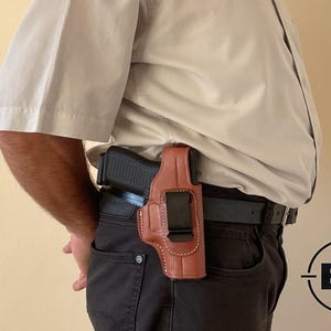 May include: A brown leather holster with a black pistol secured inside, attached to a black belt. The holster is worn on the right side, with visible stitching and a metal clip. The person is wearing a light tan button-down shirt and dark trousers.