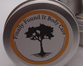 May include: A small, silver metal tin with a round, yellow and white label. The label features a black tree silhouette and the text "Finally Found It Body Care". Several similar tins are visible in the background.
