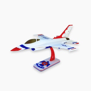 Puede incluir: Maqueta de avión blanca con detalles rojos y azules, incluido el texto "USAF" en las alas. El modelo está montado sobre un soporte con un soporte rojo y una base rectangular con un diseño rojo, blanco y azul. La cola presenta un patrón rojo y blanco.