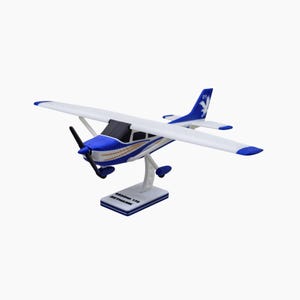 May include: A model airplane, a Cessna 172 Skyhawk, is displayed on a white stand. The plane features a white body with blue accents on the nose, tail, and wingtips. The propeller is black, and the stand has the plane's name.