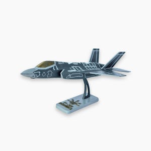 Op de afbeelding: Gedetailleerd model van een grijs en zwart F-35 straaljager, gemonteerd op een zilveren standaard. De straaljager heeft ingewikkelde paneeldetails en een geel getinte cockpit. De basis heeft tekst en een afbeelding.