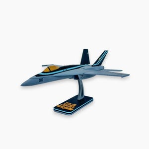May include: A model F/A-18E Super Hornet fighter jet in grey and black, with a yellow cockpit and blue detailing. It is mounted on a black stand with gold text that reads "F/A-18E Super Hornet".