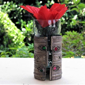May include: A silver metal vase with intricate designs and red and green gemstones. A single red flower is in the vase.