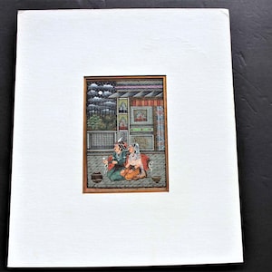 May include: A framed miniature painting depicting a couple sitting on a balcony. The painting is set against a backdrop of a building with a window and a cloudy sky. The couple is dressed in traditional Indian clothing and is holding a cup and a plate.