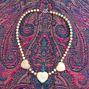 May include: A necklace made of light brown wooden beads with three heart-shaped pendants. The necklace is on a red and blue patterned fabric.