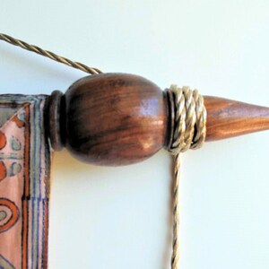 May include: Close-up of a wooden wall hanging rod with a decorative finial. The finial is a dark brown color with a gold-colored rope wrapped around it. A patterned fabric with orange and blue designs is attached.