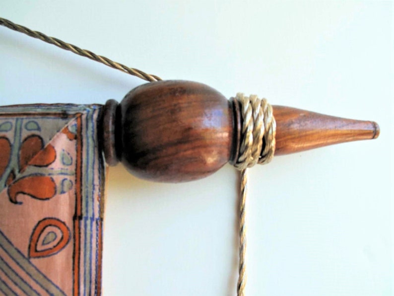 May include: Close-up of a wooden wall hanging rod with a decorative finial. The finial is a dark brown color with a gold-colored rope wrapped around it. A patterned fabric with orange and blue designs is attached.