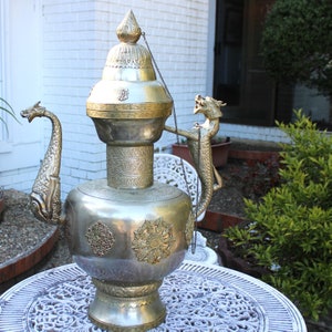 May include: A silver and gold teapot with a dragon handle and a decorative lid. The teapot is sitting on a white table with a floral pattern.