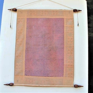 May include: A rectangular wall hanging with a pink patterned center and a tan border. The hanging is suspended by a wooden rod at the top and bottom, with decorative finials. The background is a white wall with a glimpse of a green landscape.