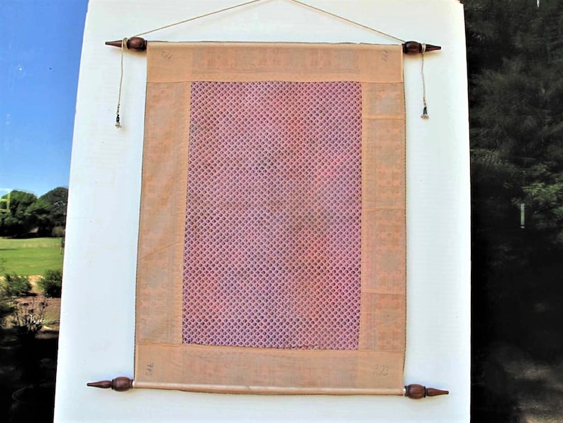 May include: A rectangular wall hanging with a pink patterned center and a tan border. The hanging is suspended by a wooden rod at the top and bottom, with decorative finials. The background is a white wall with a glimpse of a green landscape.