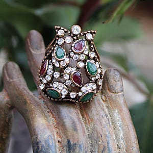 May include: A gold-toned ring with a teardrop design features a cluster of clear, red, and green gemstones. The ring is set on a wooden surface.