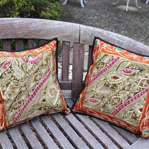 May include: Two decorative throw pillows with intricate gold, orange, and pink embroidery. The pillows feature a detailed pattern with floral and bird motifs, outlined with black trim. They rest on a wooden bench, showcasing their vibrant colors and textures.