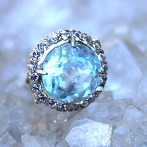 May include: A ring with a large, faceted, light blue gemstone surrounded by a halo of smaller, clear gemstones.