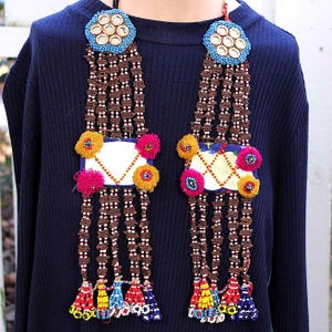 May include: Two beaded necklaces with intricate designs. Each necklace features a circular top with mirrored accents, blue beads, and multiple strands of brown beads. Colorful tassels and pom-poms adorn the lower portion, creating a vibrant and detailed accessory.