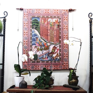 May include: A decorative tapestry with a detailed illustration of a battle scene, featuring figures on horseback and a fortress. The tapestry has a pink border with a repeating pattern and is hung on a wall. The scene is colorful and intricate.