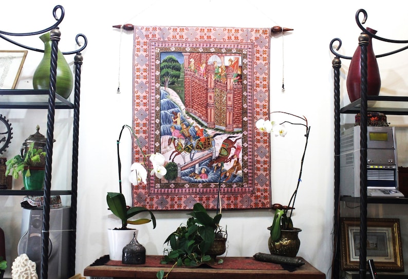 May include: A decorative tapestry with a detailed illustration of a battle scene, featuring figures on horseback and a fortress. The tapestry has a pink border with a repeating pattern and is hung on a wall. The scene is colorful and intricate.