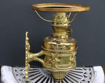 May include: A brass wall-mounted oil lamp with ornate details and a decorative shade. The lamp has a clear glass chimney and a brass burner. The lamp is labeled "Standard Oil Company".
