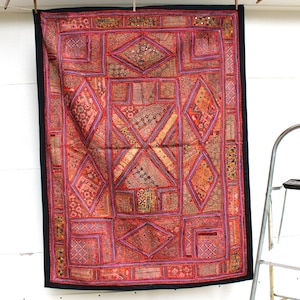 May include: A colorful patchwork tapestry with a geometric design in shades of red, pink, orange, and purple. The tapestry is framed with a black border and has a variety of textures and embellishments.