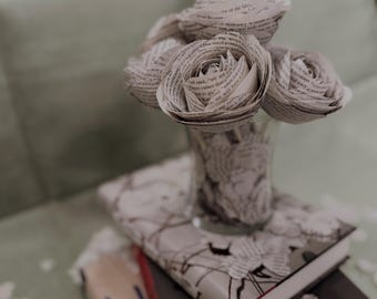 Puede incluir: Un jarrón de vidrio transparente contiene rosas de papel, apoyado sobre una pila de libros. Las rosas están hechas de páginas de texto, creando un arreglo floral único. Los libros tienen diseños florales y de texto, con el nombre del autor "SARAH J. MAAS".