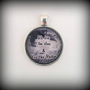 Könnte beinhalten: Silberner Anhänger mit einem Schwarzweißbild einer strukturierten Oberfläche und dem Text "We are the clay & You the Potter".