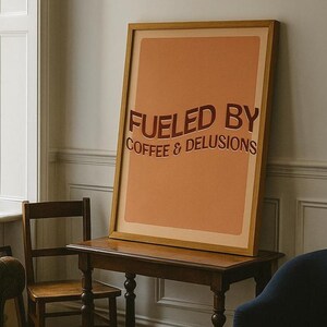 May include: A framed poster with the text "FUELED BY COFFEE & DELUSIONS" in dark red lettering. The poster has a light orange background and a wooden frame. The poster is displayed on a dark brown wooden table.