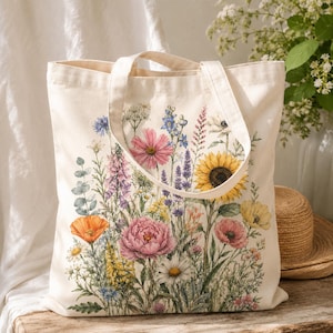 Op de afbeelding: Een crèmekleurige canvas draagtas met een levendige, gedetailleerde aquarelprint van een boeket wilde bloemen. Het bloemstuk bevat een grote gele zonnebloem, roze pioenrozen, oranje klaprozen en diverse blauwe en witte bloesems.