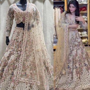 May include: A beige and pink embroidered lehenga choli set. The outfit features a long-sleeved, V-neck top and a full skirt, both heavily embellished with floral and sequin details. A sheer, matching dupatta completes the ensemble.