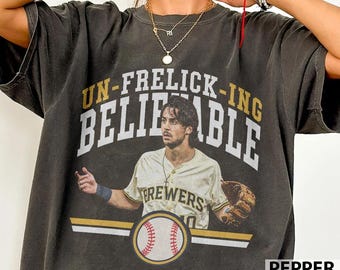 May include: A charcoal gray short-sleeve t-shirt featuring a graphic of a baseball player in a Brewers uniform with a glove. The text reads UN-FRELICK-ING BELIEVABLE above the player, with a baseball graphic and horizontal lines below.