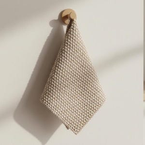 Puede incluir: Una toalla de mano beige con tejido de gofre cuelga de un gancho de pared de madera. La toalla está doblada en forma de diamante, mostrando su superficie texturizada. La pared es de color neutro claro, con una sombra.