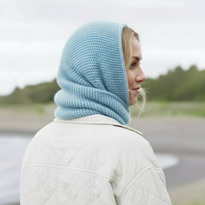 May include: A light blue knitted hood and neck warmer. The hood is pulled up over the head and neck, with the neck warmer portion visible. The person is wearing a white quilted jacket. The background shows a beach and trees.