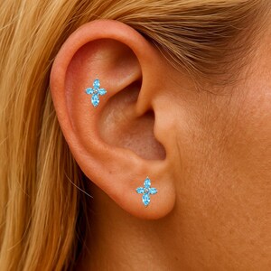 May include: Close-up of an ear adorned with two small, light blue cross-shaped earrings. Each earring features four marquise-cut gemstones surrounding a central round stone. The earrings are set against the backdrop of the ear's natural skin tone.