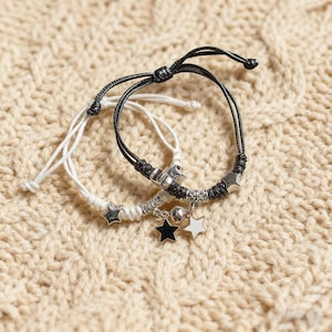 May include: Two adjustable bracelets. One is black, the other white. Both have star charms and silver-toned accents. The bracelets are on a beige, textured surface.