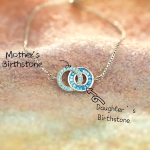 Può includere: Un braccialetto d'argento con due cerchi intrecciati, ciascuno ornato di pietre preziose blu. Un cerchio è incastonato con piccole pietre azzurre, l'altro con pietre blu rettangolari più grandi. Il testo recita "Mother's Birthstone" e "Daughter's Birthstone."