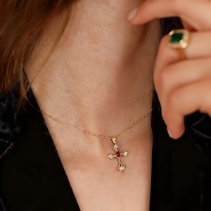 May include: A gold-colored cross pendant necklace with a small red gemstone at the center. The cross is adorned with small, clear gemstones and hangs from a delicate chain. A gold ring with a green gemstone is visible.