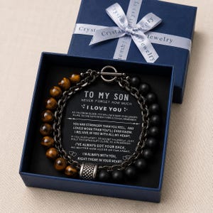 Peut inclure: Un bracelet à deux rangs avec des perles en œil de tigre et en onyx noir, présenté dans un coffret bleu avec un ruban blanc. La boîte contient une carte avec le message "To My Son".