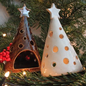 Op de afbeelding: Twee keramische kerstboomkaarsenhouders. Eén is bruin met een ster erop, de andere is wit met een ster erop. Beide hebben cirkelvormige uitsparingen. De kaarsenhouders staan op een bed van groen met rode bessen en lichtjes.