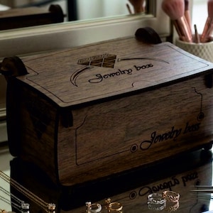 May include: A rectangular dark brown wooden box with a hinged lid, featuring a laser-engraved graphic of a diamond ring and the text Jewelry box on the top and front panels. The box has a visible wood grain texture and dark brown side accents.