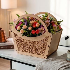 May include: A light-coloured wooden basket with a decorative laser-cut filigree pattern on the sides and two arched handles, filled with a vibrant arrangement of pink, orange, and purple flowers including roses and alstroemeria blossoms.