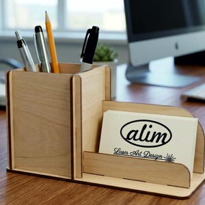 May include: A light-colored wooden desk organizer featuring a square pen holder compartment and an adjacent slot for business cards. The business card holder displays a white card with the black text alim Laser Art Design and a sun icon.