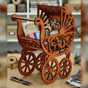 May include: A brown laser-cut wooden organizer shaped like a vintage baby carriage. The structure features intricate scrollwork patterns and four wheels. It contains a small pincushion with needles, gold-toned scissors, and thread spools.