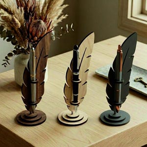 May include: Three feather-shaped pen holders made of dark wood, light wood, and black material, each standing on a circular base. Each holder features a vertical slot containing a metallic pen. The holders are arranged in a row on a wooden surface.
