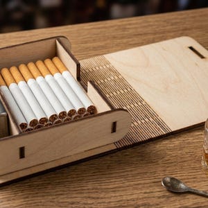 May include: A light-colored wooden cigarette case with a hinged lid and a dedicated side compartment for a metal lighter. The interior holds a row of cigarettes with orange filters. The box features laser-cut detailing and a flat, extended base.